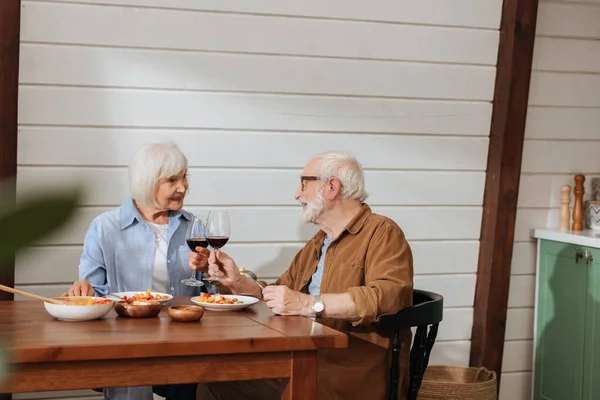 Sonriente pareja de ancianos mirándose mientras brindan con copas de vino en la mesa con cena vegetariana en la cocina - foto de stock
