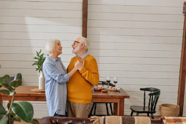 Sorrindo casal olhando um para o outro enquanto dançando juntos perto da mesa com jantar na cozinha — Fotografia de Stock