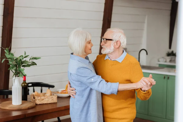 Sonriente pareja de ancianos mirándose mientras bailan cerca de la mesa con la cena sobre fondo borroso en la cocina - foto de stock