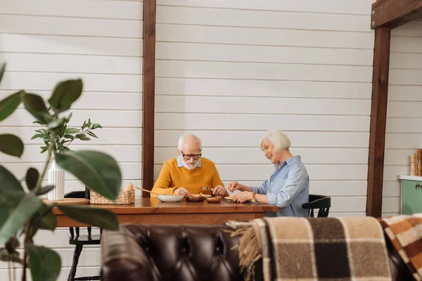 Souriant couple âgé dîner à table sur le premier plan flou dans la cuisine — Photo de stock