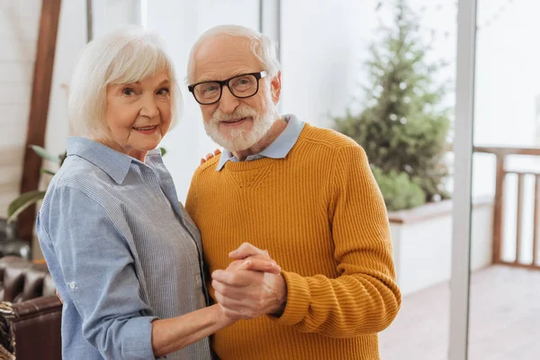 Lächelndes älteres Paar blickt in die Kamera, während es zu Hause auf verschwommenem Hintergrund gemeinsam tanzt — Stockfoto