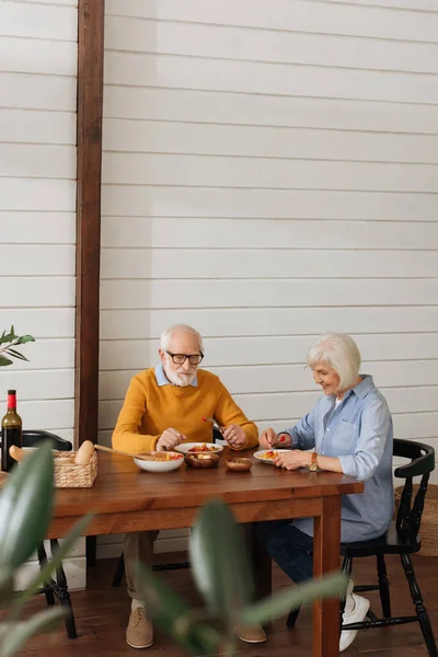Повна довжина усміхненої старшої пари їсть вечерю, сидячи за столом на кухні на розмитому передньому плані — стокове фото