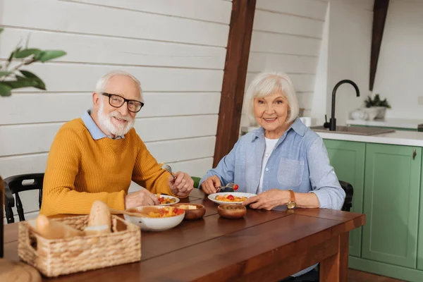 Улыбающаяся пожилая пара смотрит в камеру, сидя за столом с вегетарианским ужином на размытом переднем плане на кухне — стоковое фото