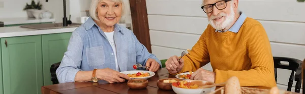 Lächelndes älteres Ehepaar, das in die Kamera blickt, während es am Tisch mit vegetarischem Abendessen auf verschwommenem Hintergrund sitzt, Banner — Stockfoto