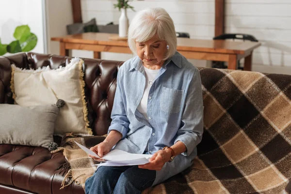 Seniorin schaut auf Scheine, während sie auf Couch vor verschwommenem Hintergrund im Wohnzimmer sitzt — Stockfoto