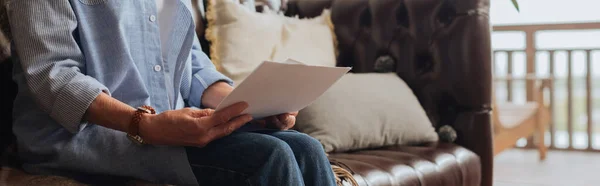 Cropped view of senior woman holding bills while sitting on couch on blurred background at home, banner — Stock Photo
