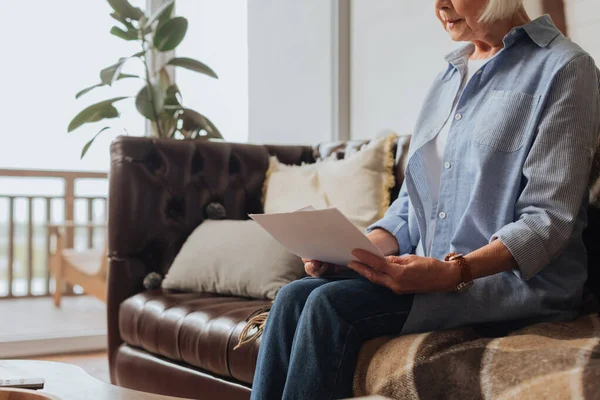 Vue recadrée de la femme âgée tenant des factures tout en étant assis sur le canapé sur fond flou à la maison — Photo de stock