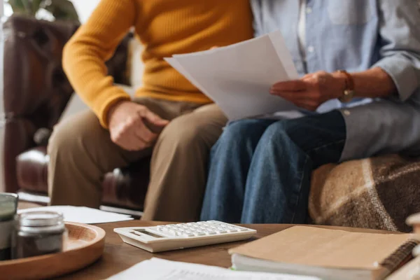 Cropped view of elderly couple with bills sitting near white calculator on table on blurred background at home — Stock Photo