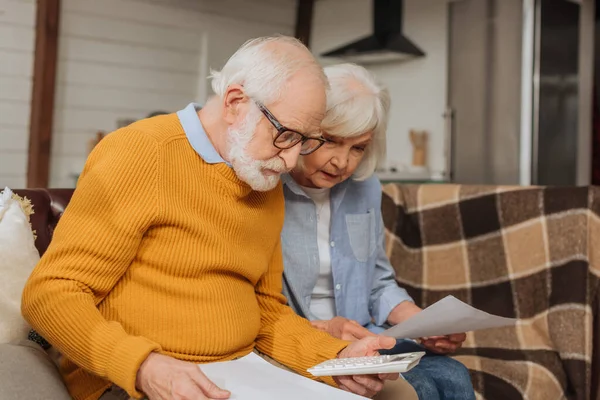 Senior couple with bills counting on calculator on couch on blurred background at home — Stock Photo