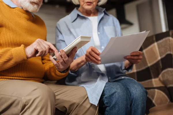 Vue recadrée du mari âgé comptant sur la calculatrice près de la femme avec des factures sur le canapé sur fond flou à la maison — Photo de stock