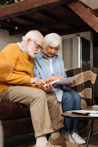 Старший муж рассчитывает на калькулятор рядом с женой глядя на счета на диване на размытом фоне в гостиной — стоковое фото