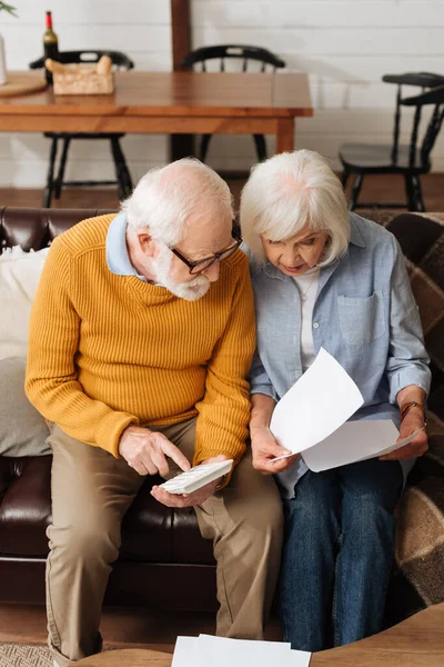 Senior Ehemann mit Taschenrechner schaut Rechnungen in der Nähe Frau auf Couch auf verschwommenem Hintergrund zu Hause — Stockfoto