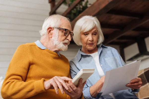 Senior Ehemann mit Taschenrechner schaut Rechnungen in der Nähe Frau auf verschwommenem Hintergrund zu Hause — Stockfoto