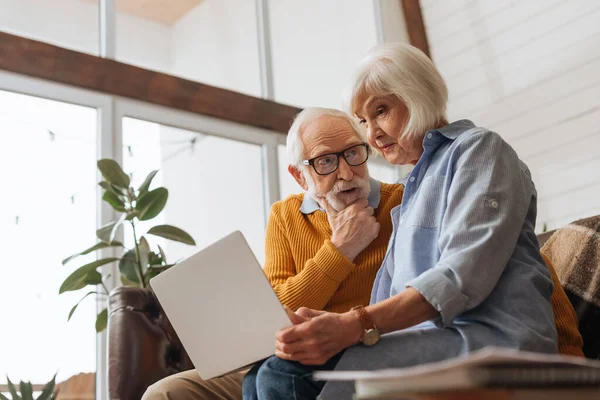 Thoughtful senior husband looking at wife with laptop on couch on blurred foreground at home — Stock Photo