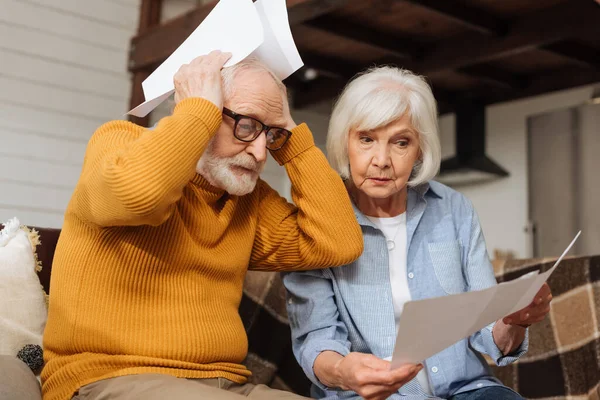 Disappointed senior husband holding bills near head sitting near wife on couch on blurred background at home — Stock Photo