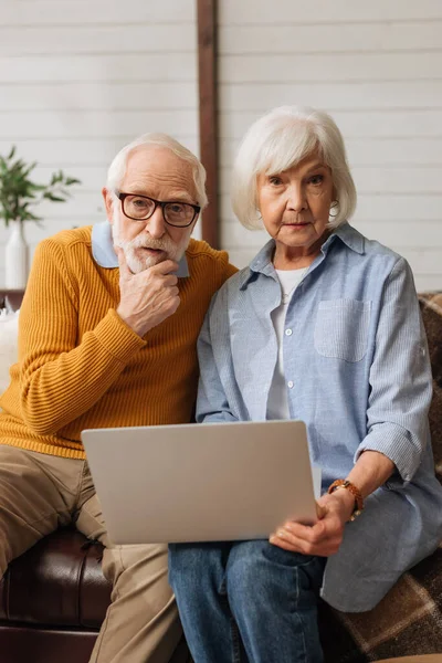 Pareja de ancianos reflexivo con portátil mirando a la cámara mientras está sentado en el sofá sobre fondo borroso en casa - foto de stock