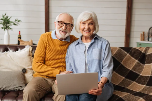 Cheerful elderly husband hugging wife with laptop while looking at camera on couch in living room on blurred background — Stock Photo