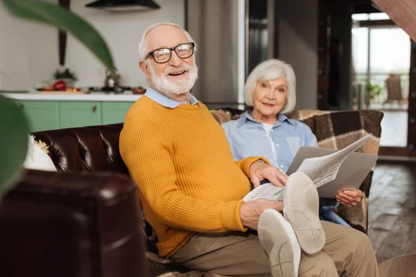 Glücklicher älterer Mann mit Zeitung blickt in Kamera nahe Ehefrau auf Couch mit verschwommener Pflanze im Vordergrund zu Hause — Stockfoto