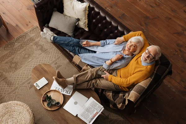 Вид сверху на пожилую пару, смотрящую в камеру, обнимаясь на диване в гостиной — стоковое фото