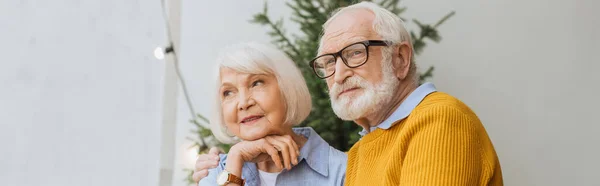 Пожилой муж смотрит в сторону, обнимая улыбающуюся жену на террасе на размытом фоне, баннер — стоковое фото