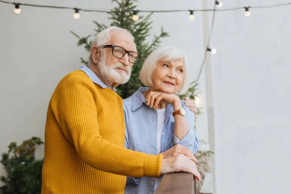 Elderly husband hugging smiling wife and looking away on terrace on blurred background — Stock Photo