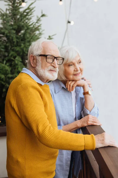 Senior Ehemann umarmt lächelnde Frau, während er auf der Terrasse vor verschwommenem Hintergrund wegschaut — Stockfoto