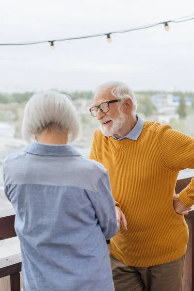 Back view of elderly wife standing near smiling husband with hand on hip on terrace on blurred background — Stock Photo