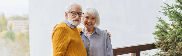 Lächelndes älteres Paar blickt in die Kamera, während es sich auf der Terrasse vor verschwommenem Hintergrund umarmt, Banner — Stockfoto