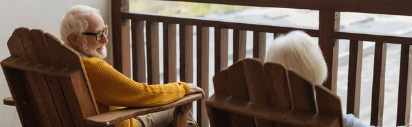 Smiling senior husband looking at wife in armchair on terrace on blurred background, banner — Stock Photo