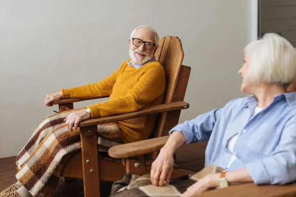 Sonriente anciano mirando a su esposa mientras está sentado en un sillón de madera con manta a cuadros en la terraza en primer plano borroso - foto de stock