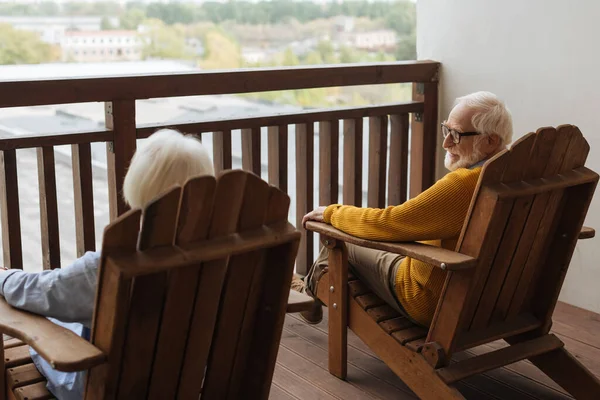 Vista posteriore della moglie con i capelli grigi seduta vicino al marito sorridente in poltrona di legno su sfondo sfocato sulla terrazza — Foto stock