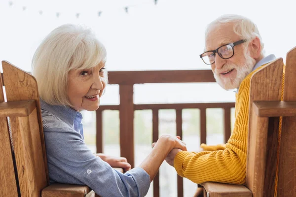 Smiling elderly couple looking at camera and holding hands while sitting in armchairs on terrace on blurred background — Stock Photo