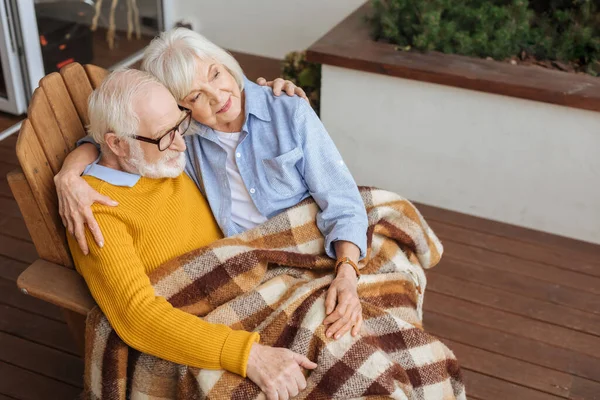 Sorridente coppia anziana con coperta a quadri, abbracciare e distogliere lo sguardo mentre seduto in poltrona sulla terrazza su sfondo sfocato — Foto stock