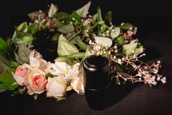 Rose bouquet and urn with ashes on black background, funeral concept — Stock Photo