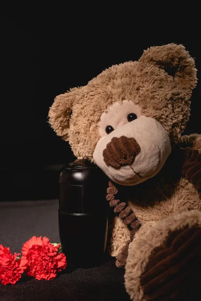 Carnation, teddy bear and urn with ashes on black background, funeral concept — Stock Photo