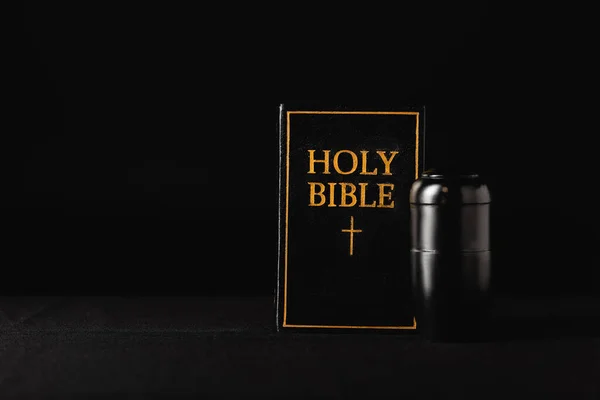 Heilige Bibel und Urne mit Asche auf schwarzem Hintergrund, Bestattungskonzept — Stockfoto