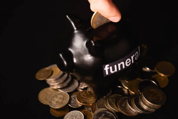 Vista recortada del hombre poniendo moneda en alcancía sobre fondo negro, concepto de funeral - foto de stock