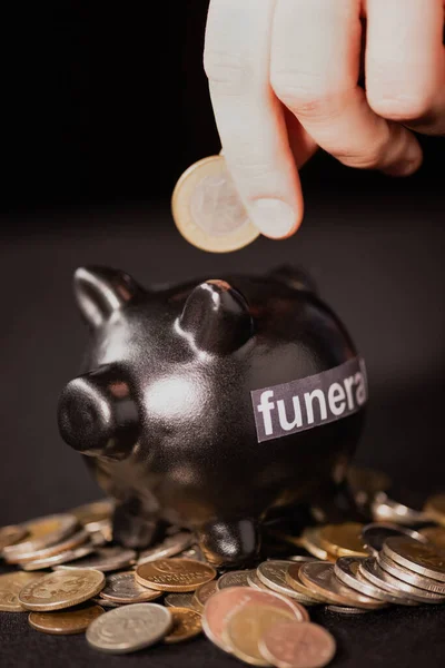 Vista recortada del hombre poniendo moneda en alcancía sobre fondo negro, concepto de funeral - foto de stock