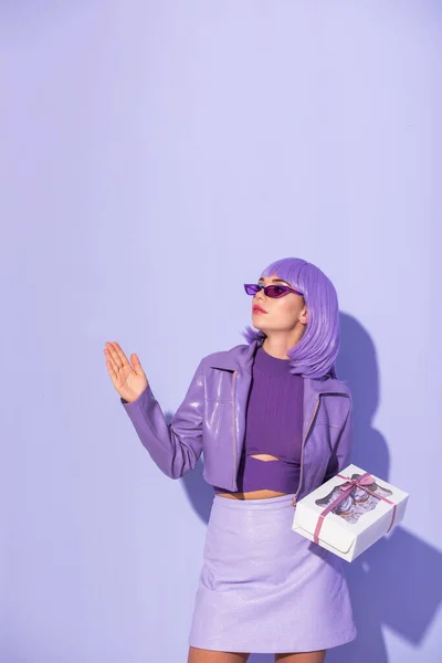 Mujer joven vestida con estilo de muñeca con caja de magdalenas sobre fondo de color violeta - foto de stock