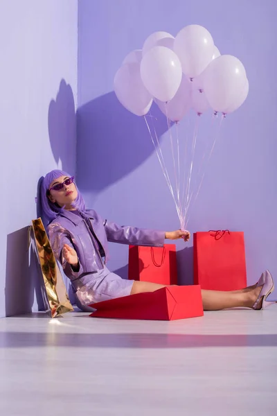 Giovane donna vestita in stile bambola seduta con borse della spesa e palloncini su sfondo viola colorato — Foto stock