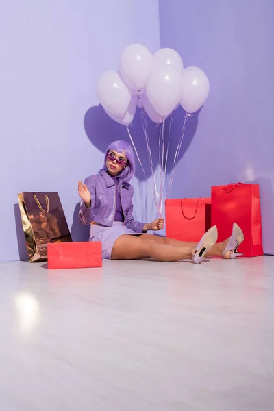 Mujer joven vestida con estilo de muñeca sentada con bolsas de compras y globos sobre fondo de color violeta - foto de stock