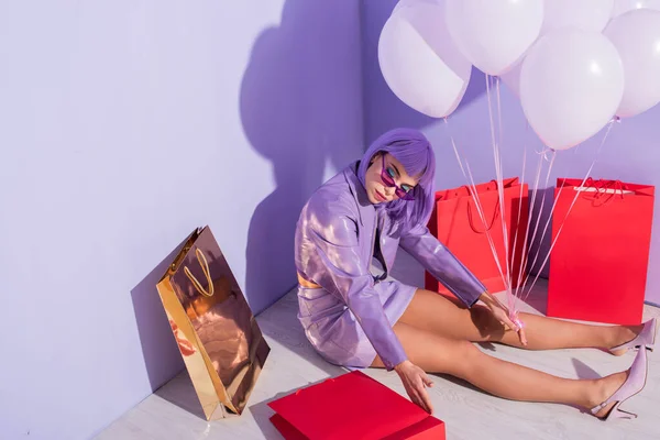 Молодая женщина одета в кукольный стиль сидя с пакетами покупок и воздушными шарами на фиолетовом красочном фоне — стоковое фото