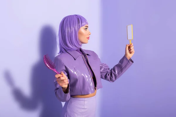 Jeune femme habillée de style poupée avec brosse à cheveux et miroir sur fond violet coloré — Photo de stock