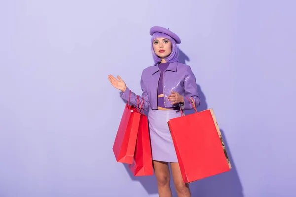 Mujer joven vestida con estilo de muñeca con bolsas de compras rojas sobre fondo de color violeta — Stock Photo