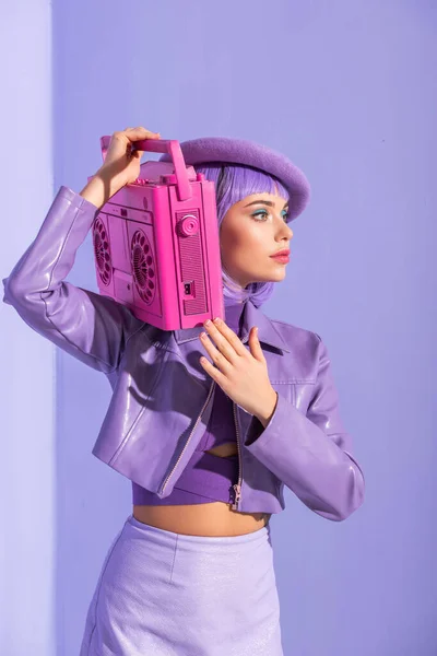 Mujer joven vestida con estilo muñeca posando con grabadora de cinta retro rosa sobre fondo colorido violeta - foto de stock