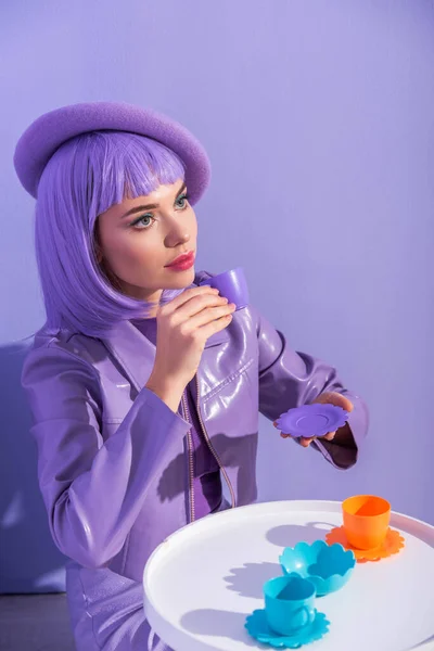 Junge Frau im Puppenstil in Baskenmütze posiert mit Spielzeuggeschirr auf violettem Hintergrund — Stockfoto