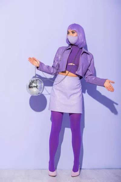 Jeune femme habillée de style poupée en masque médical avec boule disco sur fond violet coloré — Photo de stock