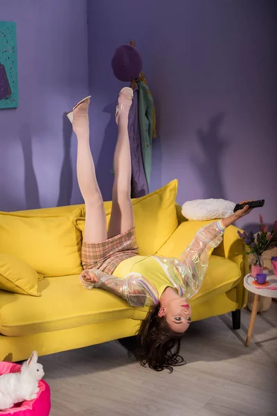 Young woman posing as doll on yellow couch with remote control — Stock Photo