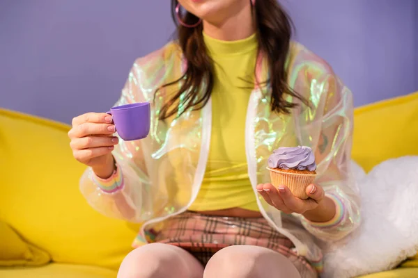 Vista recortada de la joven mujer haciéndose pasar por muñeca con cupcake y taza de juguete en el sofá amarillo - foto de stock