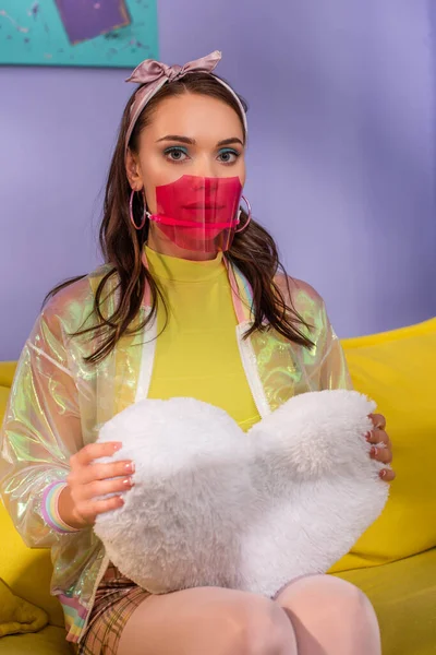 Young woman in medical mask posing as doll on yellow couch with pillow — Stock Photo